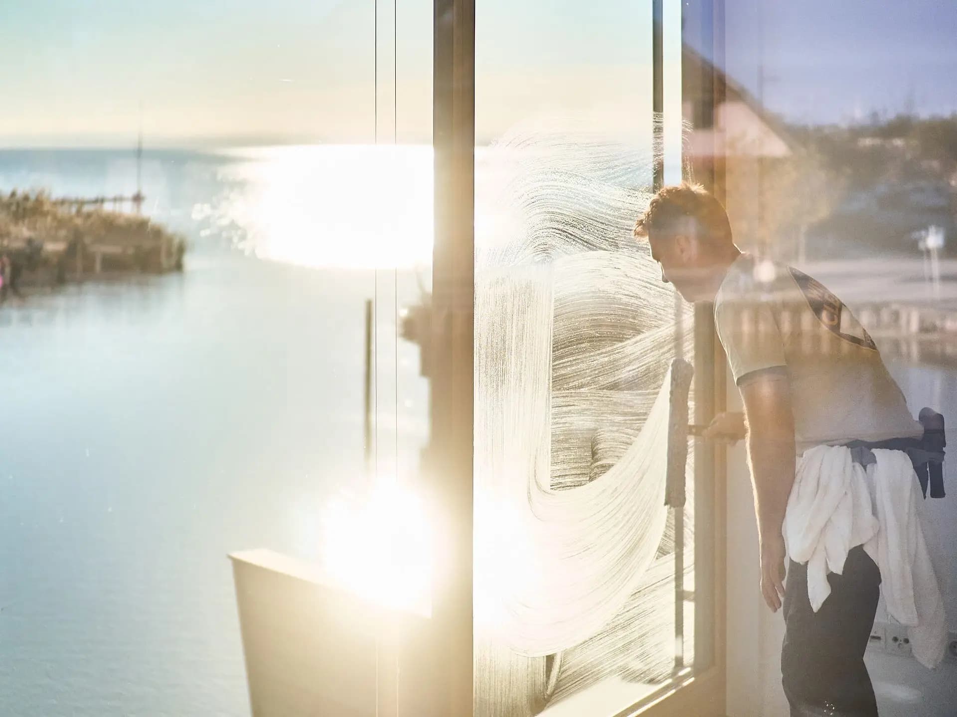 Justin sweeping soap water across a tall window at golden hour, a harbour and open water visible through the glass